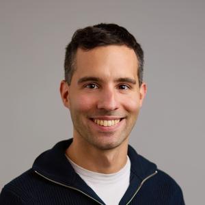 Head-and-shoulders portrait of a smiling man in a dark zip-up sweater against a plain gray background.